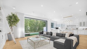 A modern living room with a large sectional sofa, a glass coffee table, and a view of a pool and greenery outside. - Spaulding Ave Farmhouse - Bespoke LA Home w/ Pool & Home Theater (Los Angeles)