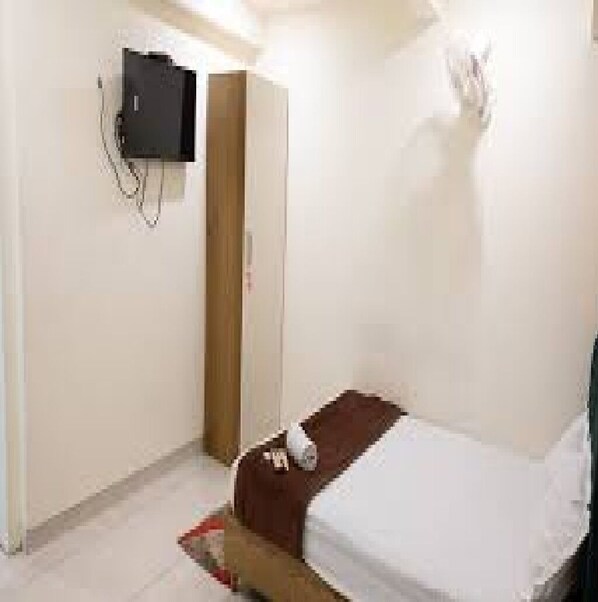A small, sparsely furnished room with a single bed, a wooden wardrobe, and a wall-mounted television. - Manbhar Heavens Hotel (Amer)