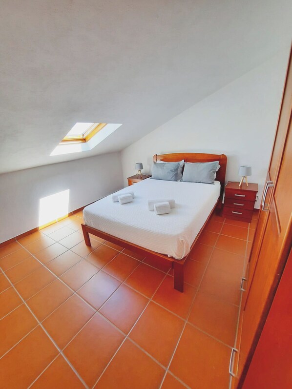 Un dormitorio con cama de madera, dos mesitas de noche, tragaluz y suelo de baldosas.