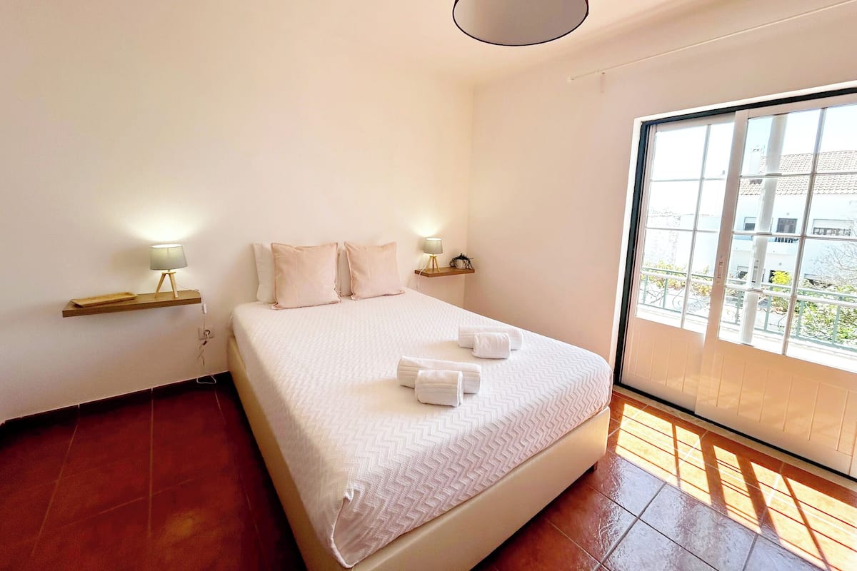 Un dormitorio con una cama grande, dos mesitas de noche con lámparas, una ventana con vistas a edificios y una lámpara de techo roja.