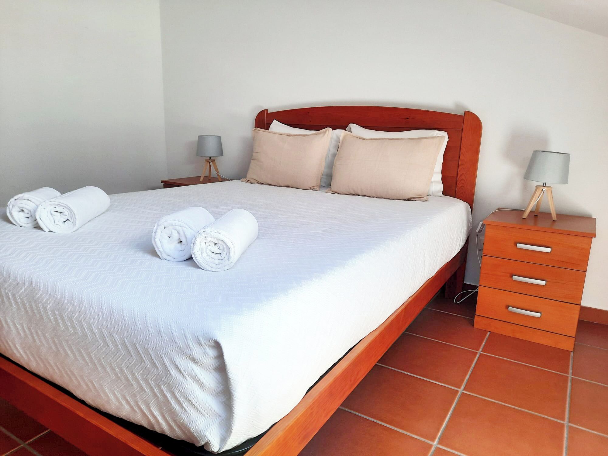 Una cama bien hecha con toallas blancas enrolladas, un cabecero de madera y una mesita de noche con lámpara.