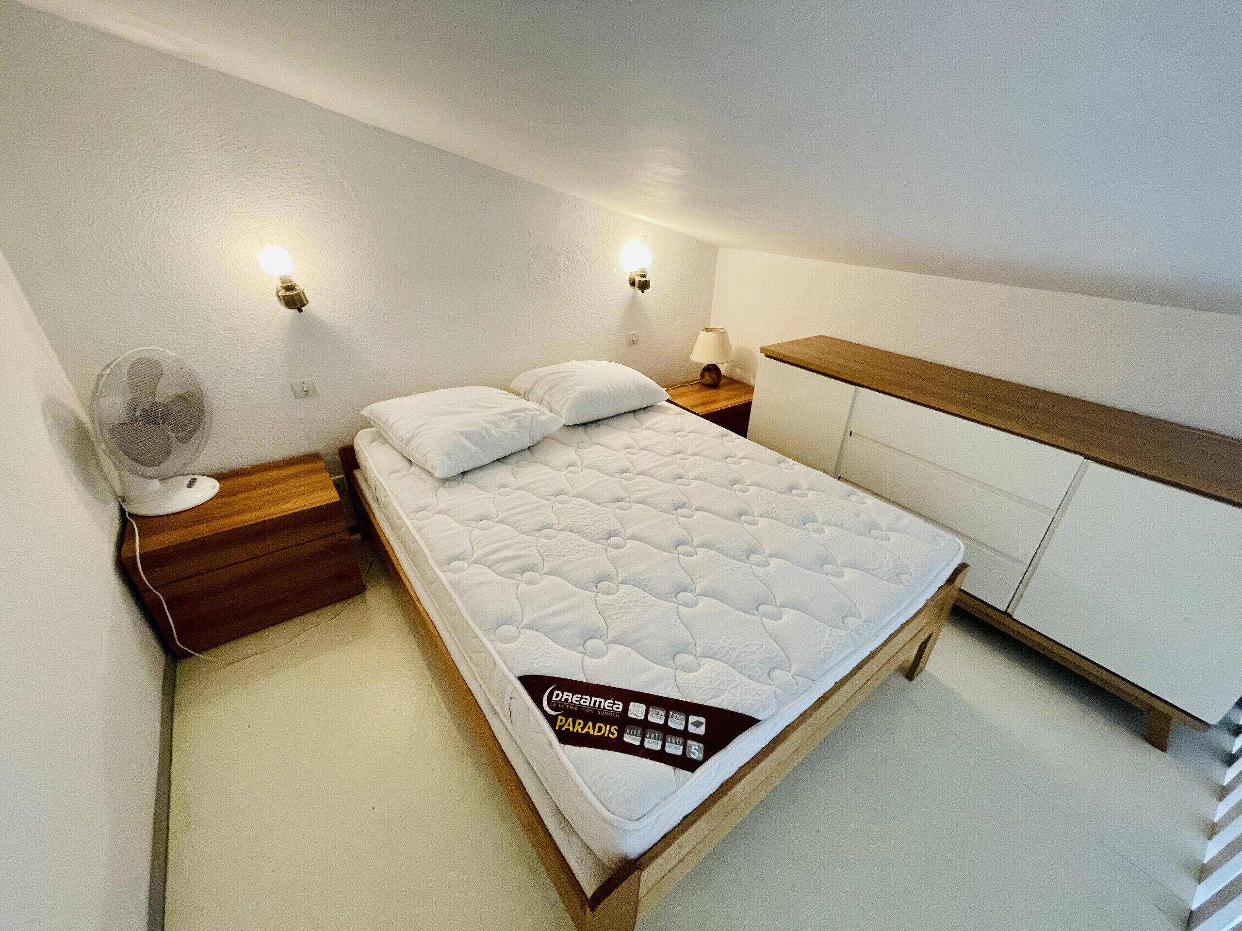A neatly arranged bedroom with a bed, bedside lamps, a nightstand, and a wall-mounted fan.