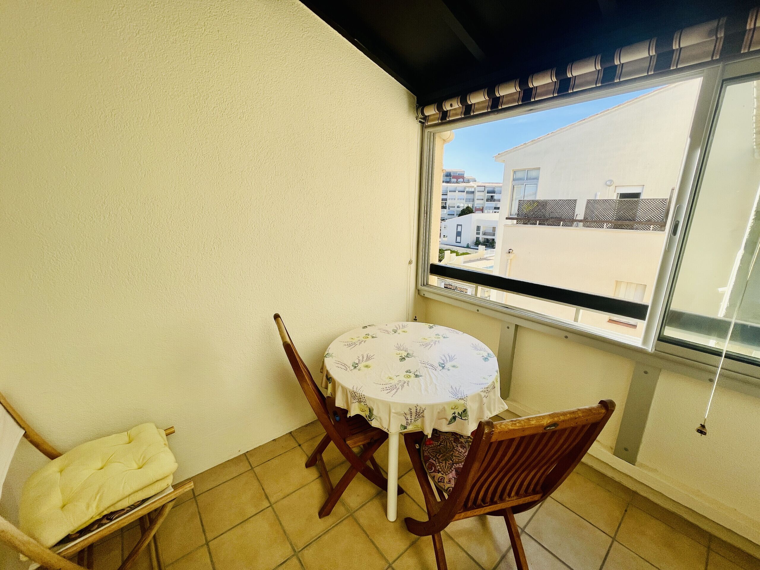 Uma pequena varanda com mesa e cadeiras, uma janela com vista para edifícios e uma cadeira com almofada.