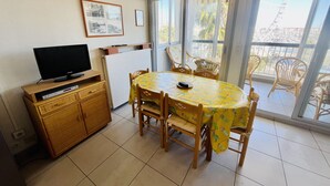 Ein Essbereich mit rundem Tisch und Stühlen, einem Fernseher auf einem Schrank und Blick ins Freie durch eine Glastür.