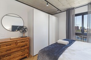 Ein Schlafzimmer mit einem hölzernen Schminktisch, einem Bett mit blauer Decke, einem Standventilator, einem großen Spiegel und Blick auf eine Baustelle durch ein Fenster.