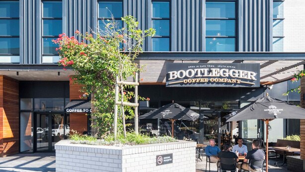 An outdoor seating area with tables and chairs in front of a building with a sign that reads "Bootlegger Coffee Company". - Central Century City Comfort | Balcony and Wi-Fi (Cape Town)