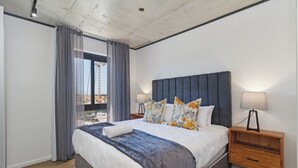 A modern bedroom with a large bed, a headboard, two bedside lamps, a wooden nightstand, and a view of the city through a window. - Central Century City Comfort | Balcony and Wi-Fi (Cape Town)