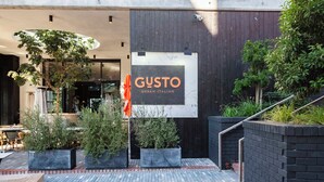 An outdoor seating area with a sign for 'GUSTO Urban Italian' restaurant, surrounded by potted plants and a brick pathway. - Central Century City Comfort | Balcony and Wi-Fi (Cape Town)