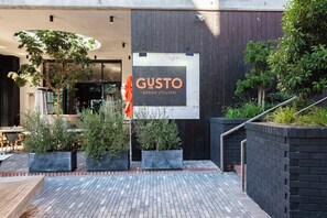 Ein Sitzbereich im Freien mit einem Schild für das Restaurant 'GUSTO Urban Italian', umgeben von Topfpflanzen und einem Pflasterweg.