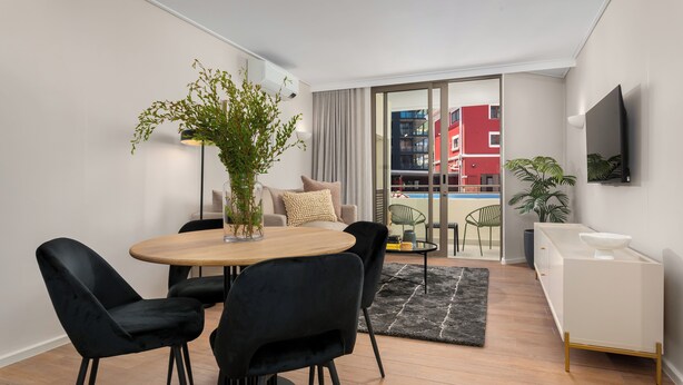 A modern living room with a dining area, a sofa set, a TV mounted on the wall, and a view of a red building through the glass door. - Dolce Vita | Stylish Sea Point Gem (Cape Town)