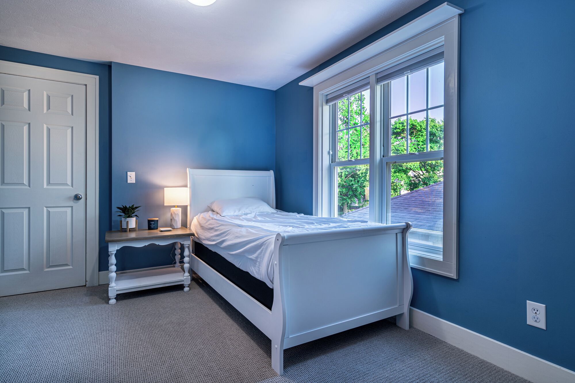 A bedroom with a single bed, a nightstand, a lamp, and a window with a view of greenery.
