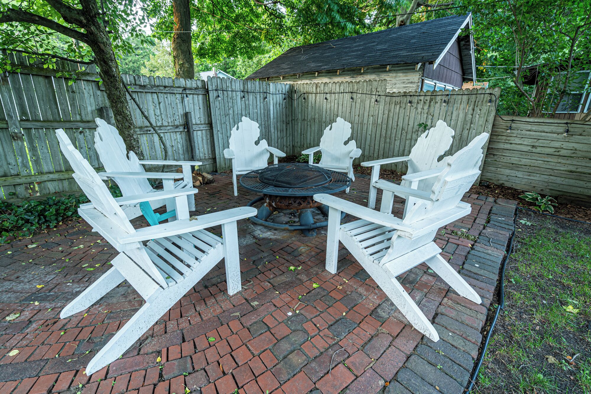 Une terrasse dotée de chaises Adirondack blanches et d’une table ronde, entourée d’une clôture en bois et d’un chemin pavé de briques.