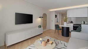 Ein modernes Wohnzimmer mit einem Flachbildfernseher, einem runden Couchtisch mit Kerzen und einer Couch. Im Hintergrund ist eine Küchenzeile mit weißen Schränken und einem Esstisch erkennbar.