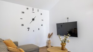 Een moderne woonkamer met een grote wandklok, een flatscreen televisie, een grijze bank en een houten salontafel met een bloemenvaas.
