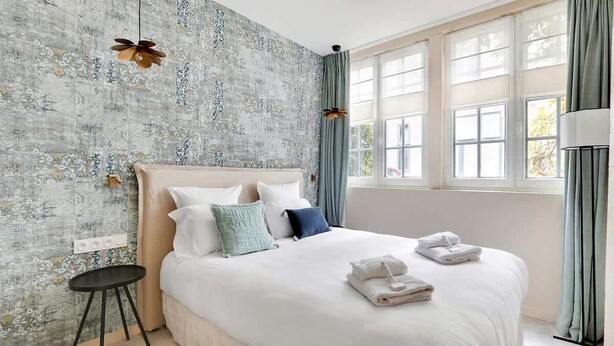 Ein ordentlich bezogenes Bett mit Kissen und einem kleinen Tisch daneben, ein Fenster mit Vorhängen sowie eine Wand mit einer strukturierten Oberfläche.