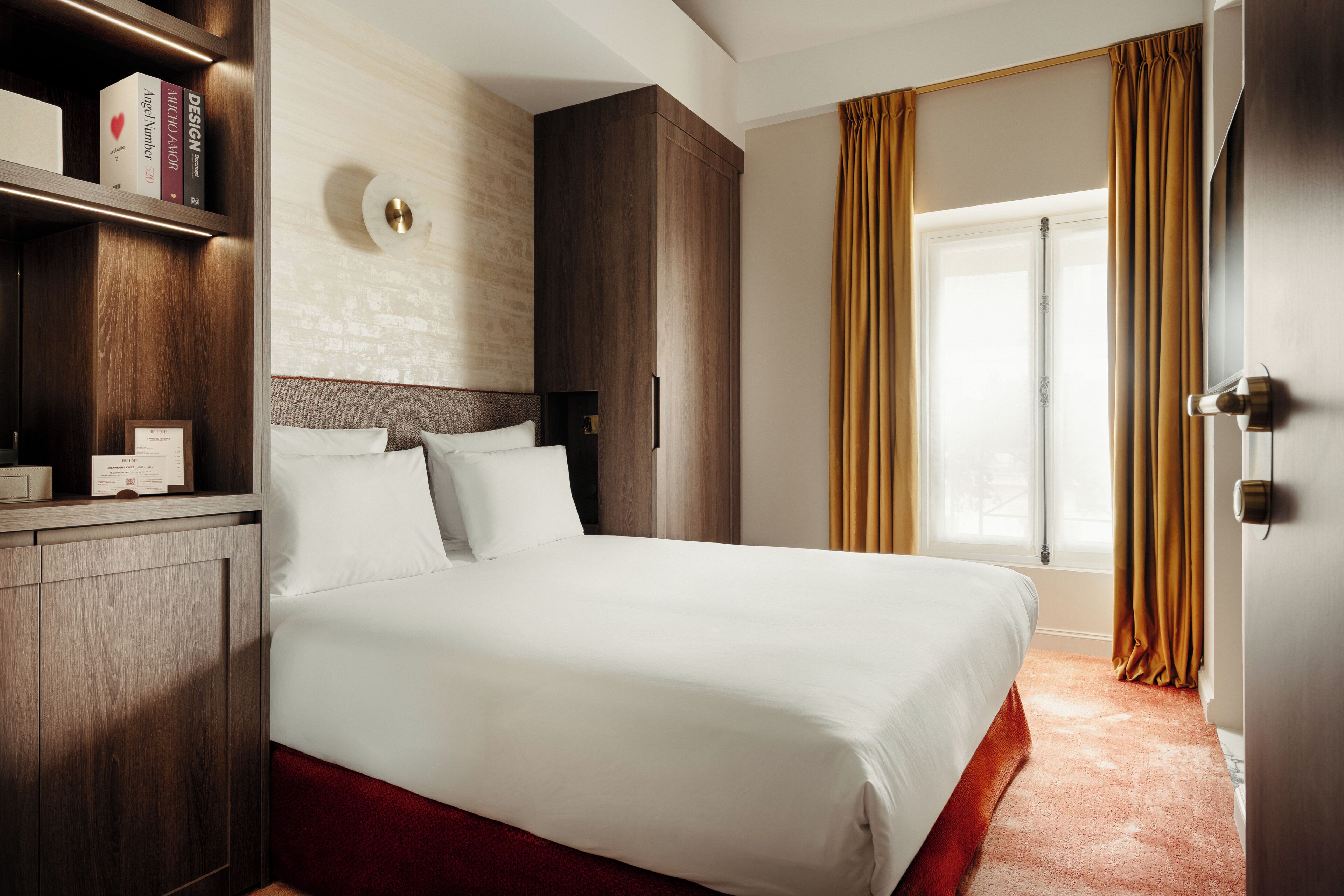 A hotel room with a large bed, wooden wardrobe, a bedside table, a wall-mounted light, and a window with curtains.