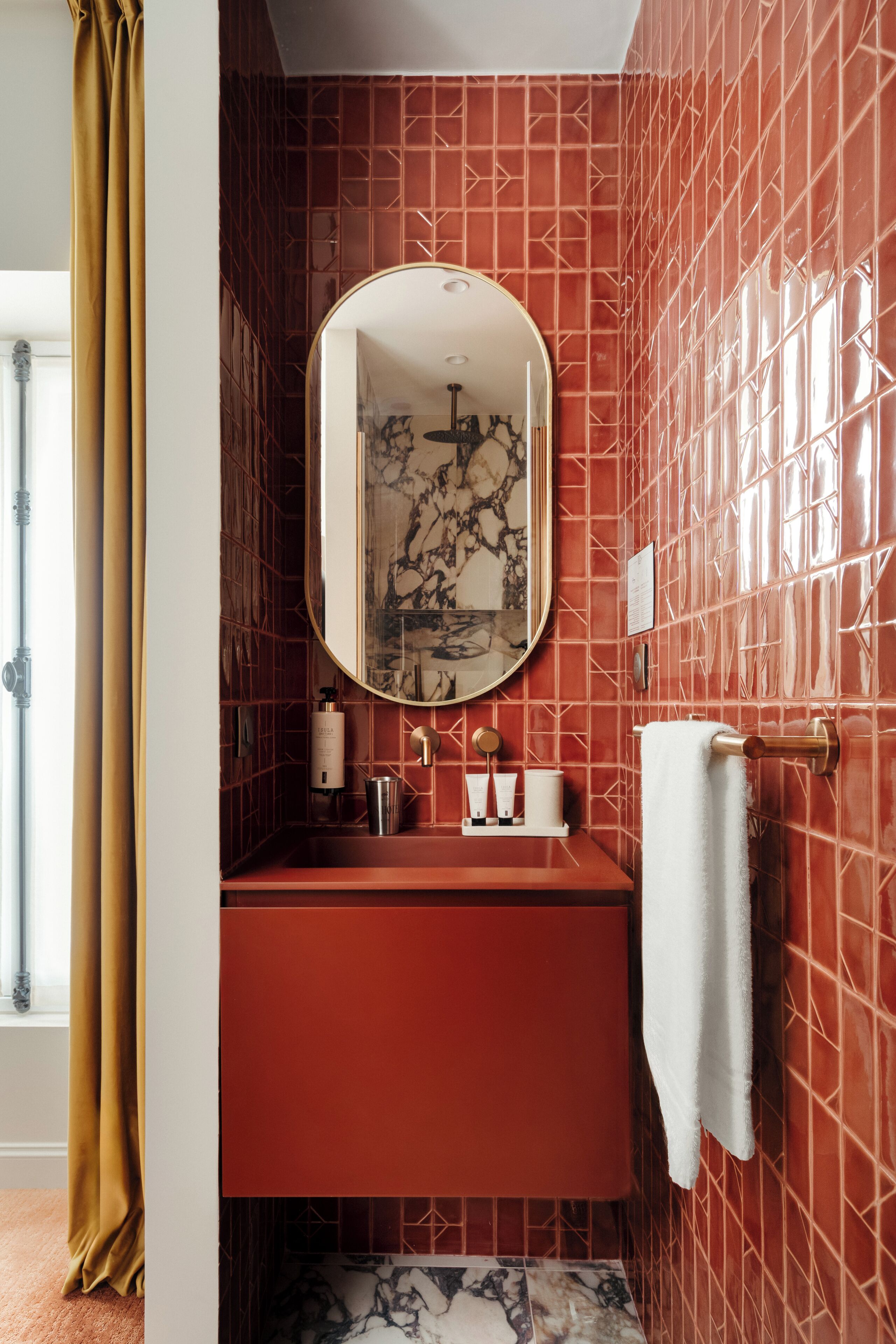 Un baño con pared de azulejos rojos, un lavamanos de mármol, un espejo y un perchero para toallas.