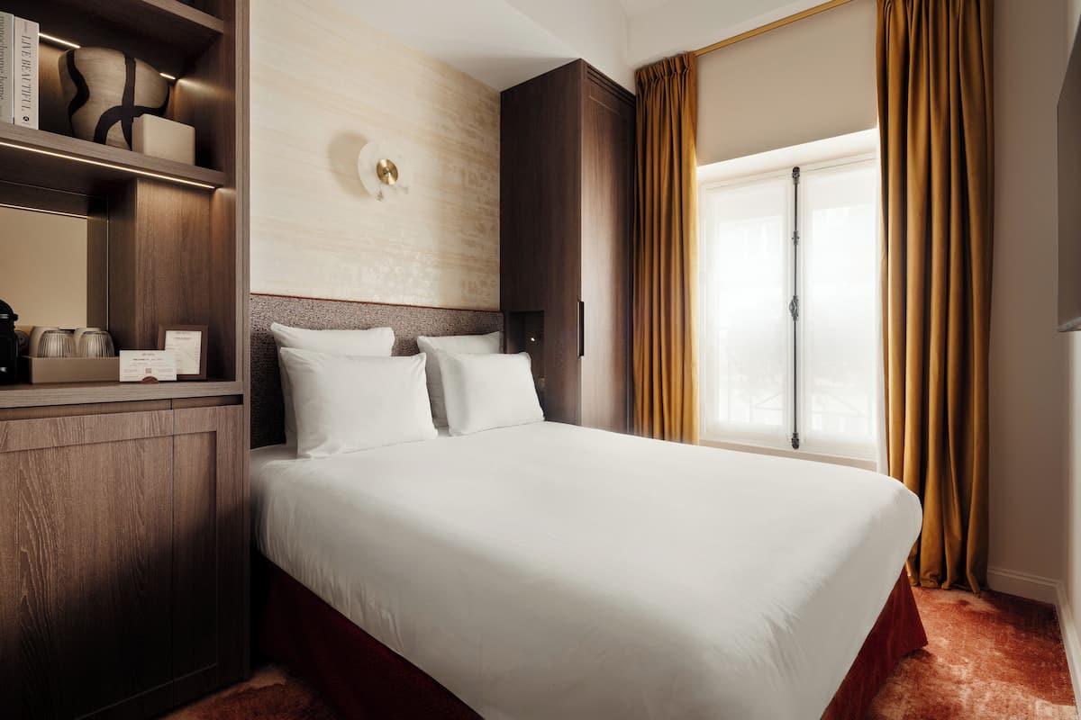 A hotel room with a large bed, wooden headboard, a bedside table with a lamp, and a window with curtains.