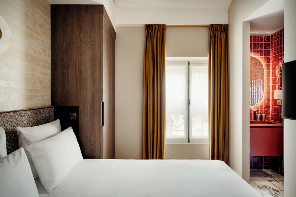 A hotel room with a bed, a wooden headboard, a window with curtains, and a red bathroom visible through the door.
