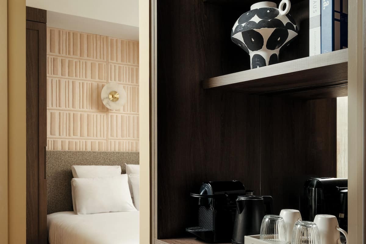A hotel room with a bed, a coffee station, a bookshelf, and a decorative wall panel.