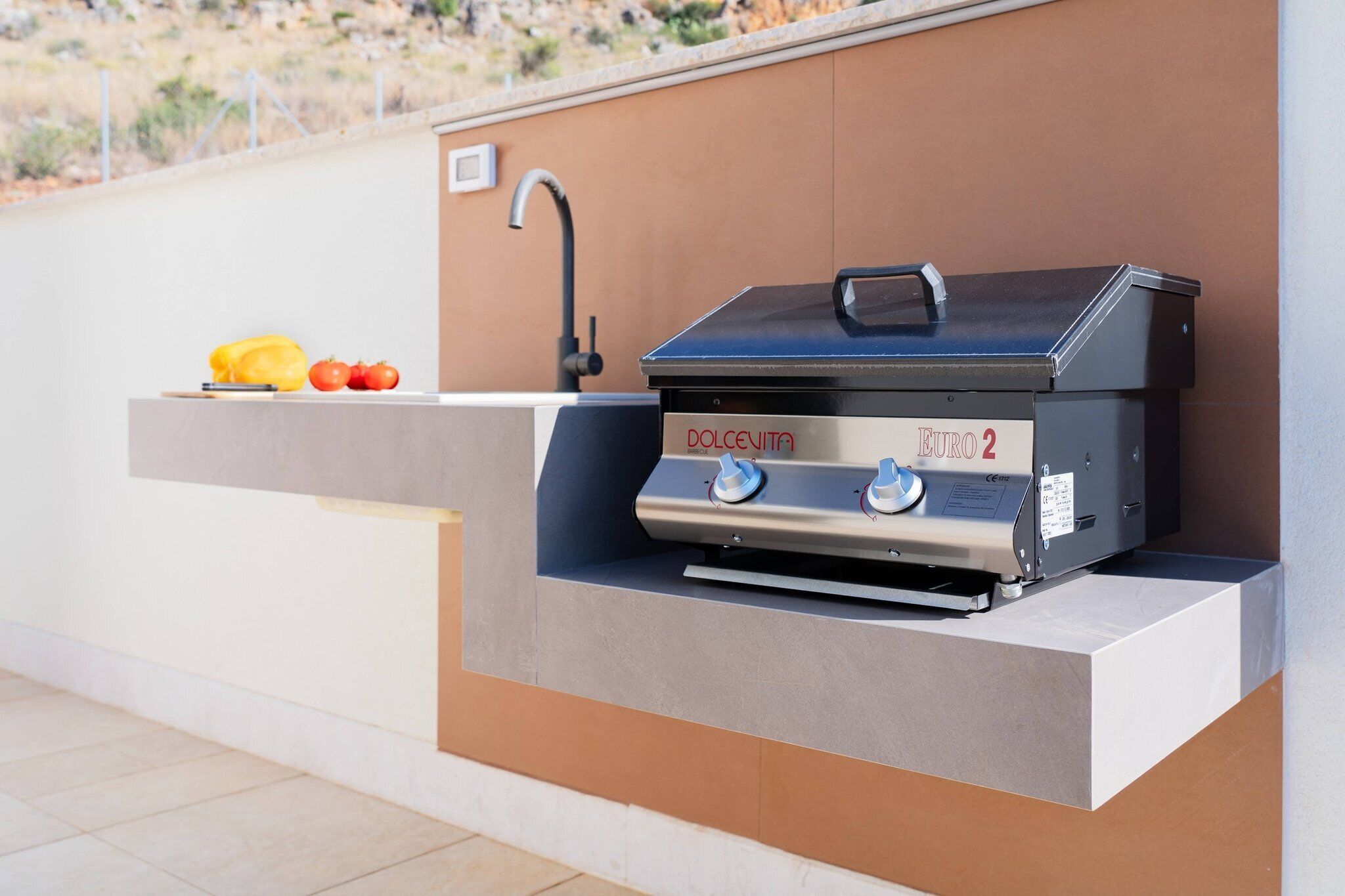 Une cuisine extérieure équipée d’un barbecue intégré, d’un évier et d’un plan de travail où sont disposés des légumes.
