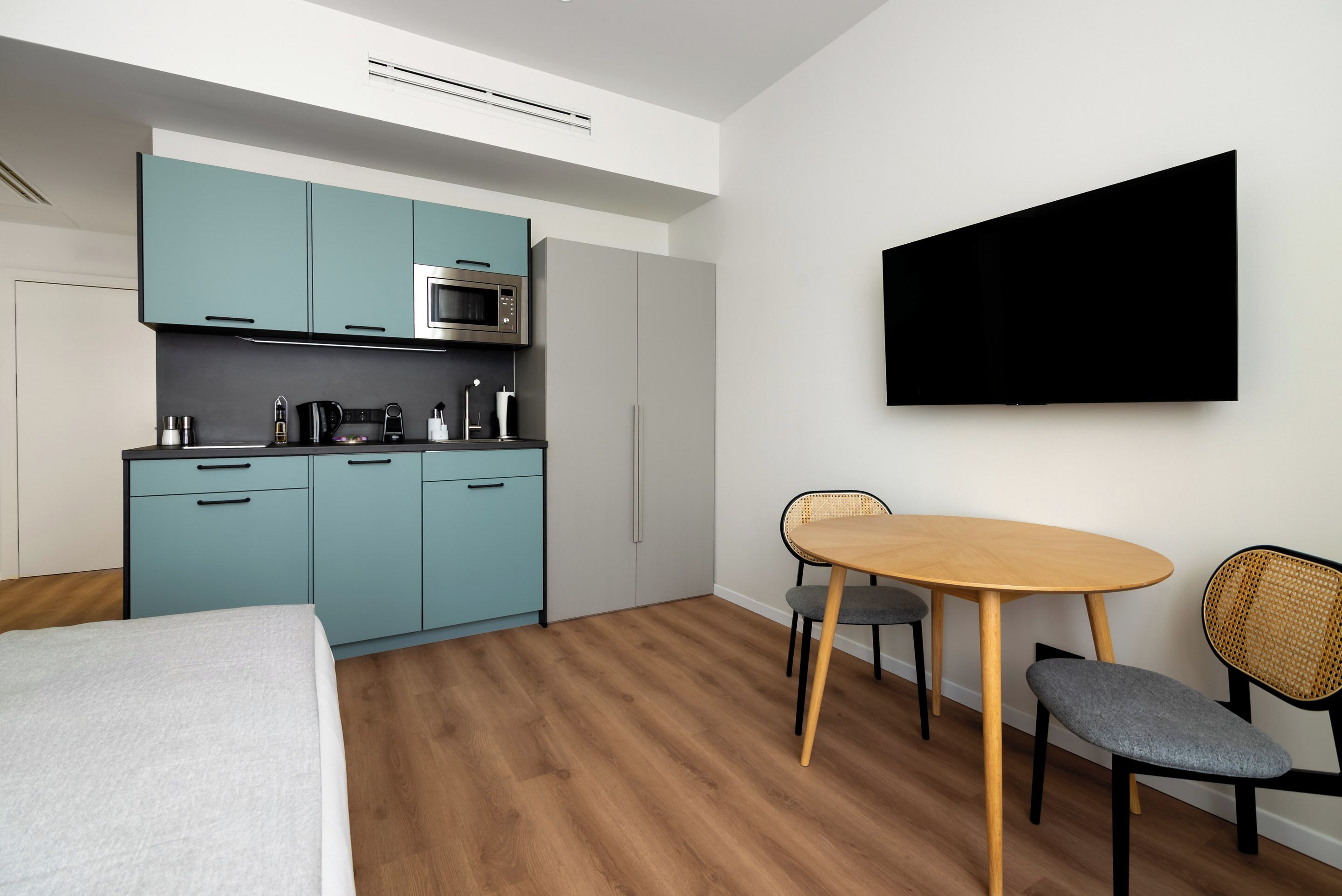 A modern kitchen with teal cabinets, a microwave, and a coffee machine. There is a dining area with a wooden table and chairs.