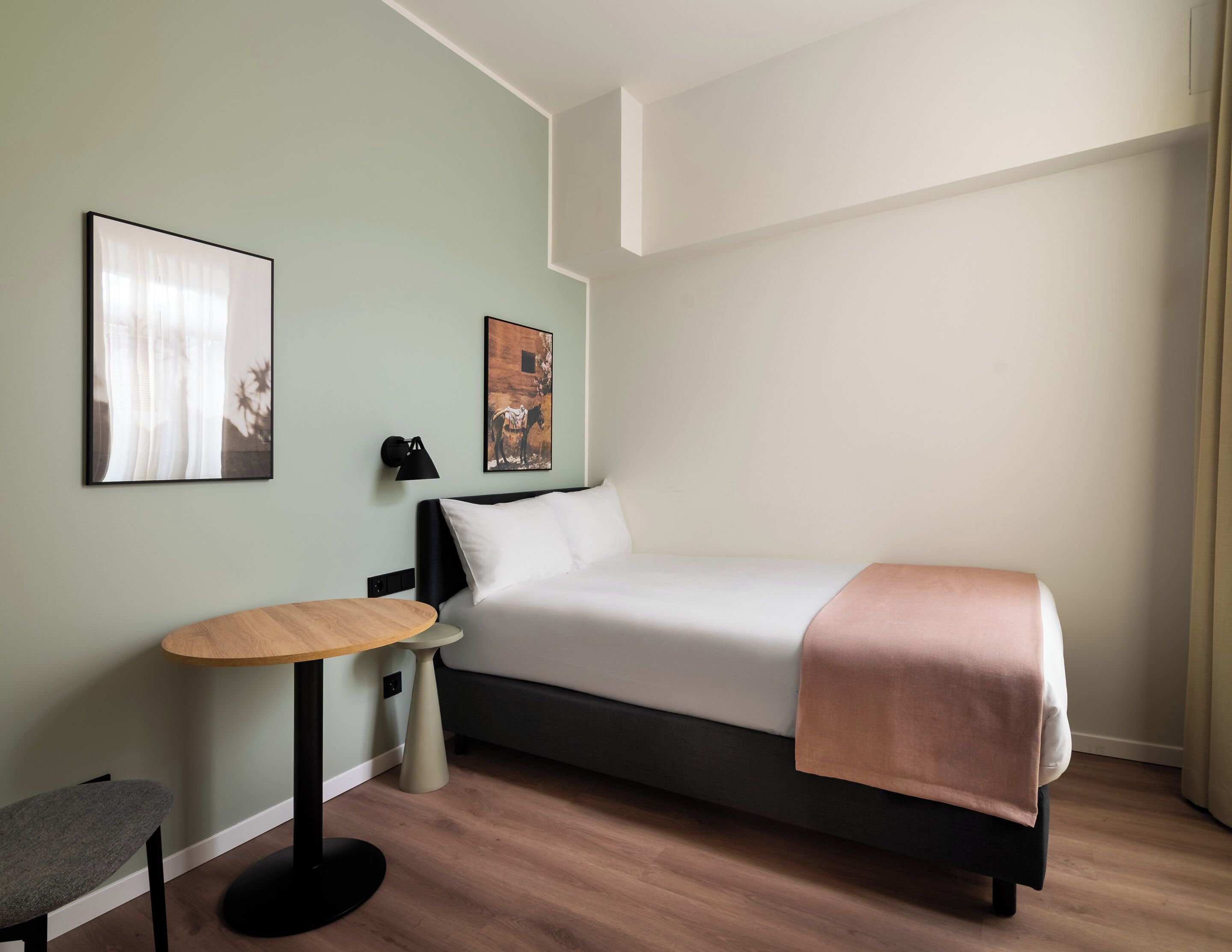 A modern hotel room with a single bed, a small round table, a chair, and two wall-mounted artworks.