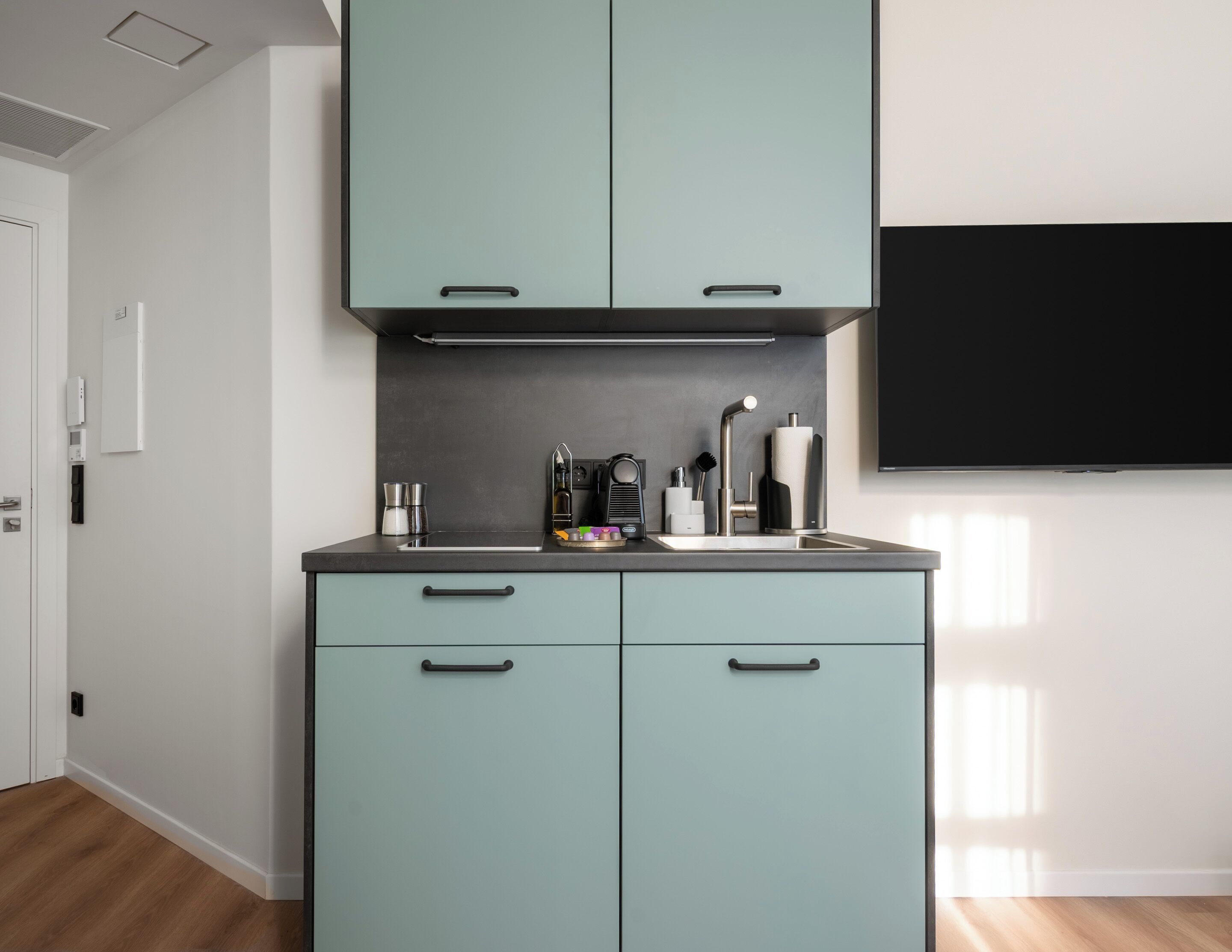 A modern kitchen with teal cabinets, a stainless steel sink, and a flat-screen TV mounted on the wall.