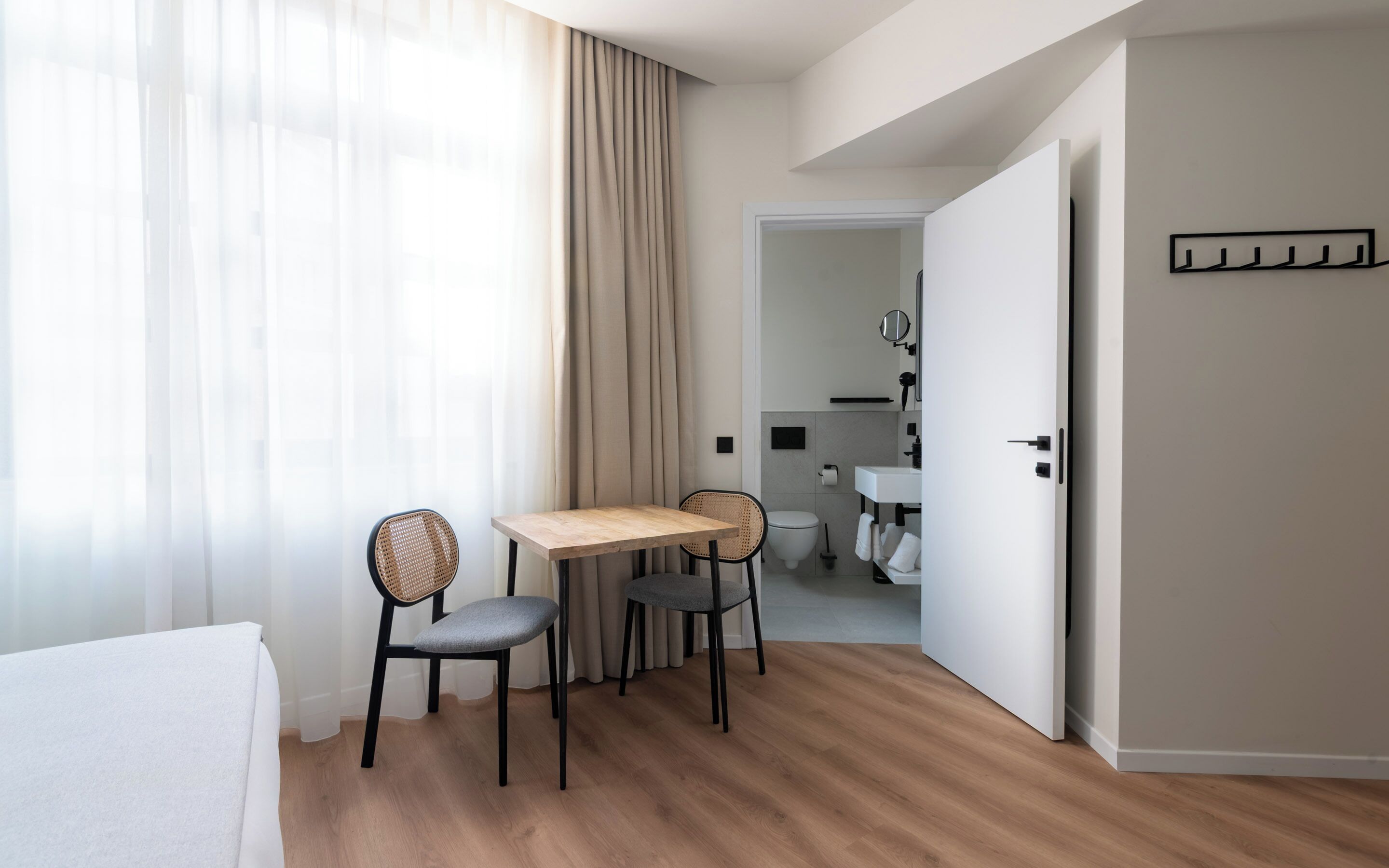 A hotel room with a wooden floor, a bed, a table, two chairs, a bathroom with a sink and toilet visible through an open door.