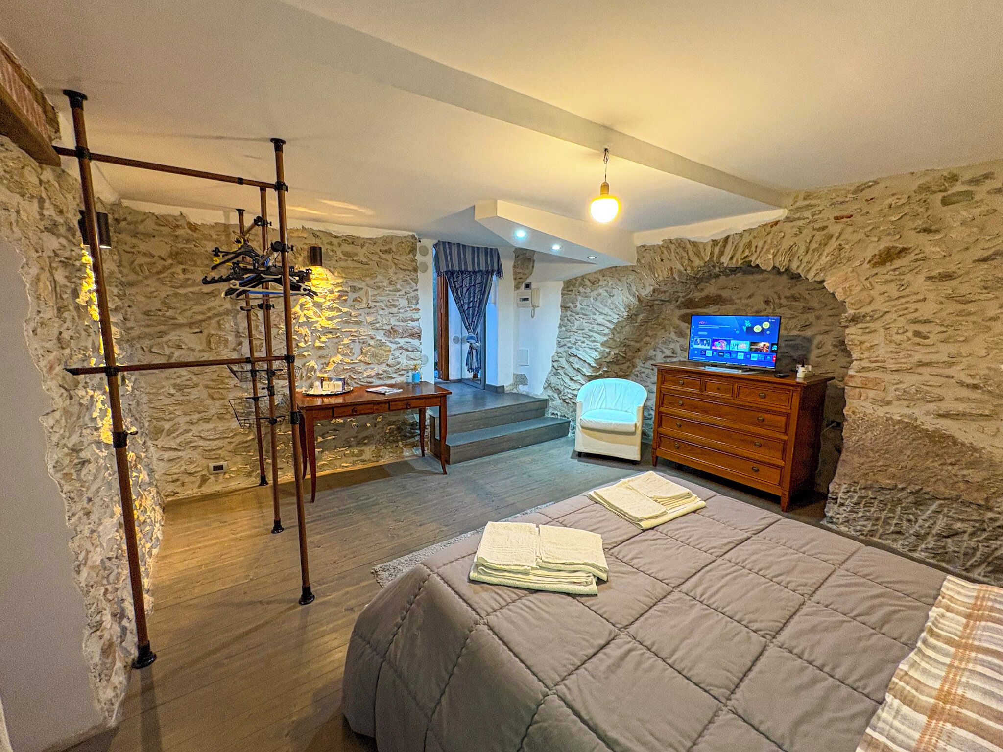 A room with a stone wall, a bed with a quilt, a wooden dresser, and a television.