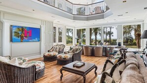 A spacious living room with wicker furniture, a large TV, and a balcony with a view of palm trees. - MyStay at 14 Laughing Gull – Premier Oceanfront Estate in Sea Pines (Hilton Head Island)