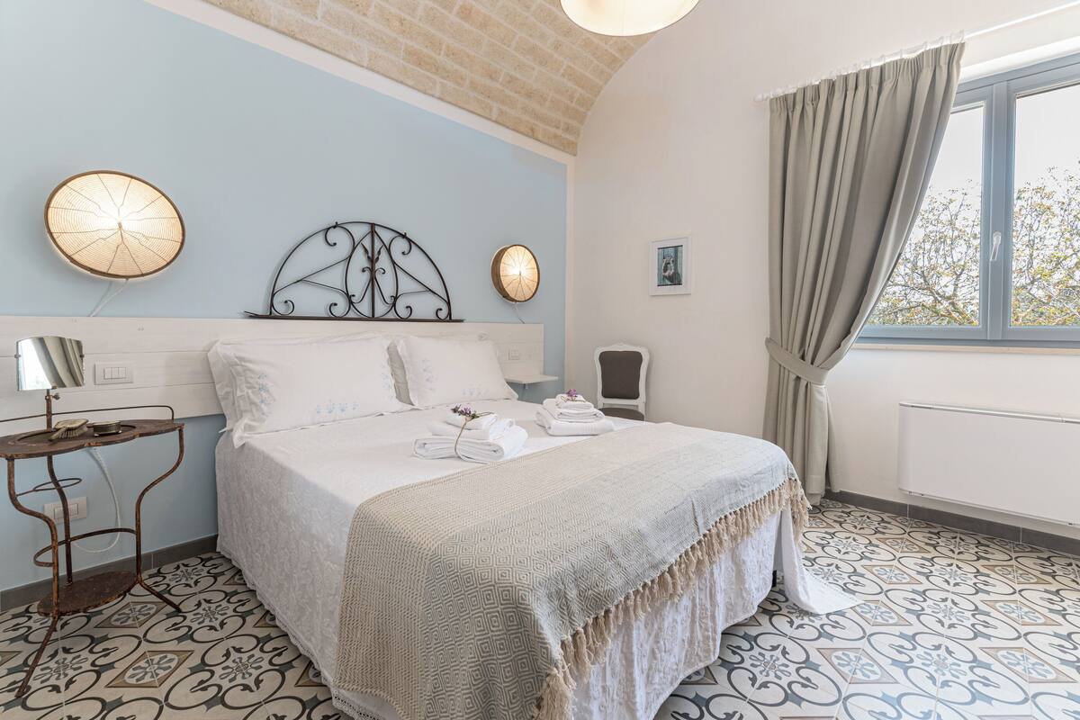 Een slaapkamer met een bed, een nachtkastje met een lamp, een wandklok en een raam met gordijnen.