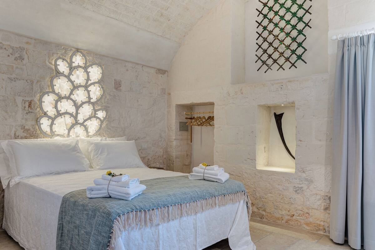 Een slaapkamer met een bed, een raam met decoratieve tralies en een klein raam met een zwart object.