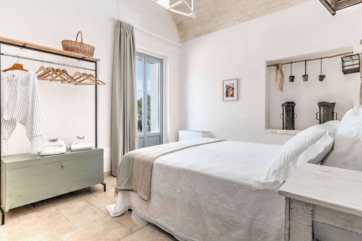Een slaapkamer met een bed, een kaptafel, een raam met gordijnen en een wandplank met een mandje.
