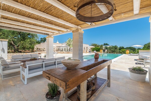 An outdoor seating area with a pool view, featuring a wooden table, white sofas, and potted plants. - Borgo Altobello (Cisternino)