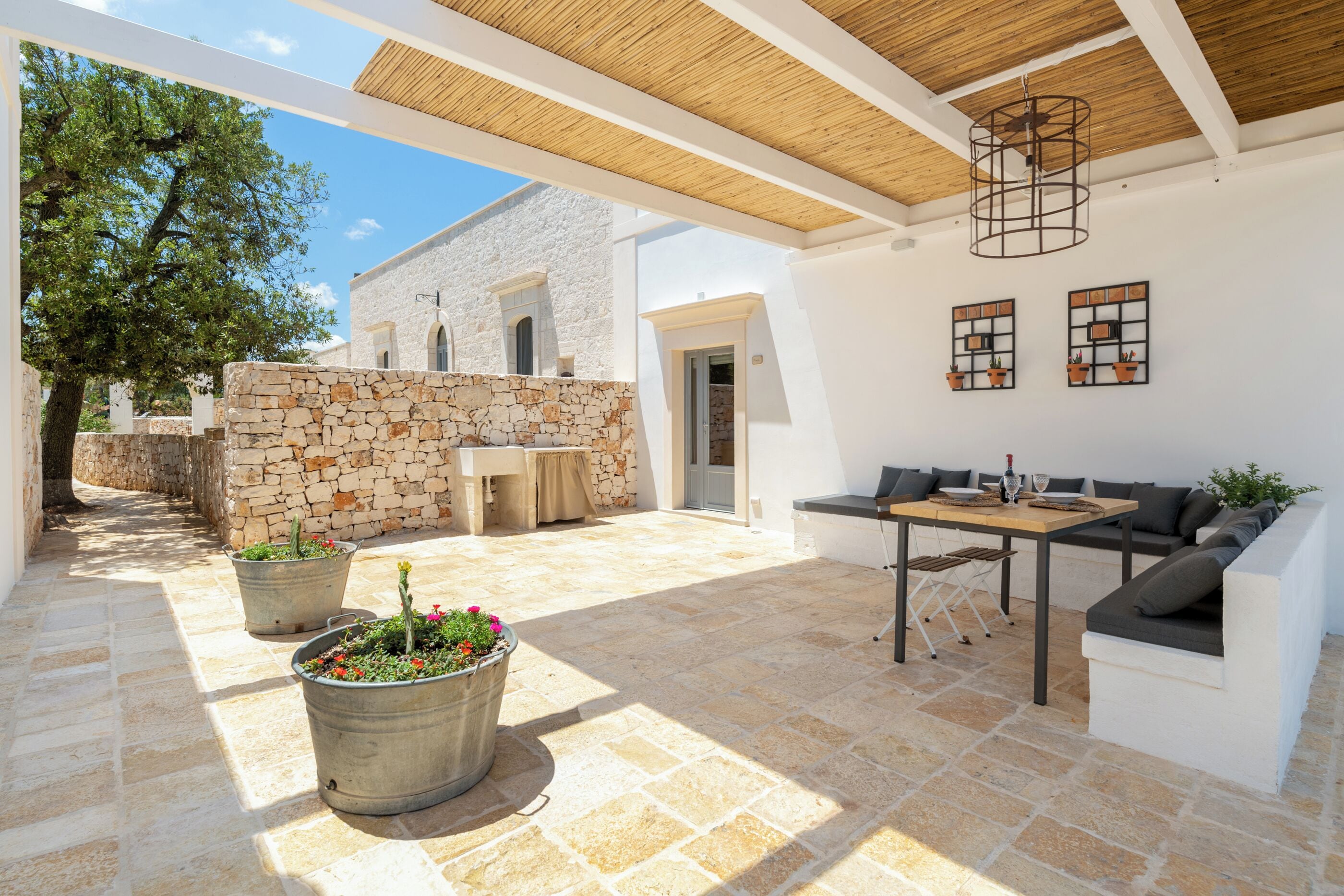 An outdoor patio with stone flooring, a wooden ceiling, and a stone wall. There is a table and a sofa set, and two potted plants.