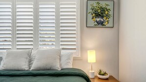 Un dormitorio con cama, almohadas, una mesita de noche con lámpara, una planta pequeña y un cuadro en la pared.