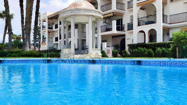 A swimming pool with a white gazebo in the center, surrounded by palm trees and a multi-story building with balconies. - Laguna Green Apartment (El Morche)