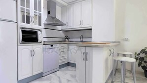 A modern kitchen with white cabinetry, a built-in oven, a microwave, and a stainless steel range hood. There are two white bar stools and a potted plant. - Laguna Green Apartment (El Morche)