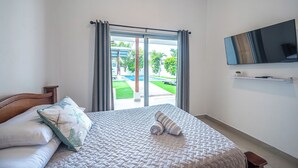 A bedroom with a bed, a TV mounted on the wall, and a view of a pool and greenery outside. - Casa Alegria Villa -Jaco Beach Great Location (Jacó)