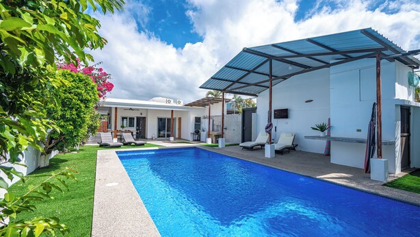 A swimming pool with a covered patio area, surrounded by a well-maintained lawn and a house with a white exterior. - Casa Alegria Villa -Jaco Beach Great Location (Jacó)