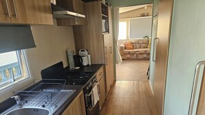 A compact kitchen with a stove, sink, and wooden cabinets. A window provides natural light, and a sofa is visible in the adjacent living area. - Seaside Escape at Nab Tower Lane (Chichester)