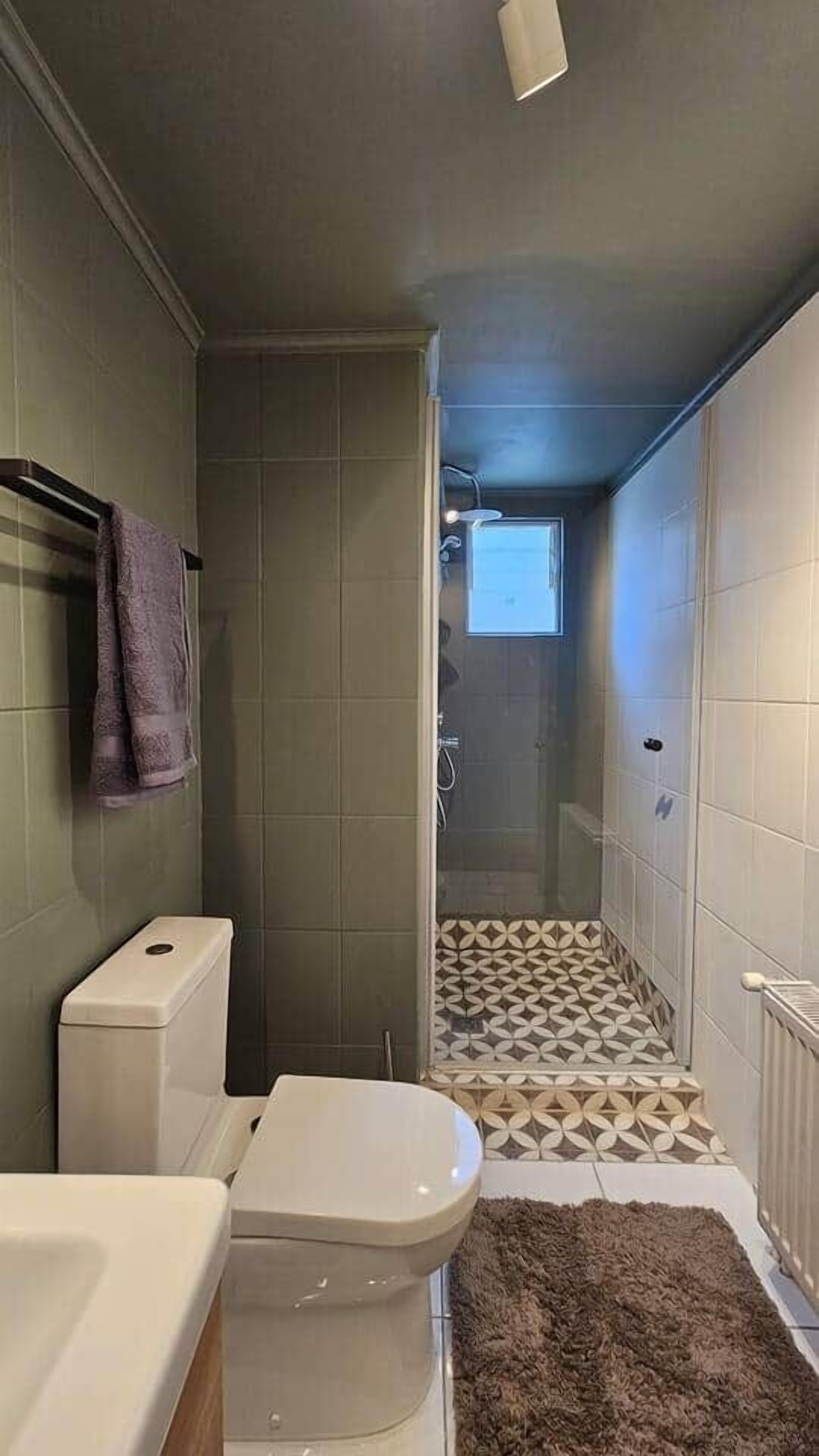 Un baño pequeño con inodoro, zona de ducha con azulejos estampados y una alfombra marrón en el suelo.