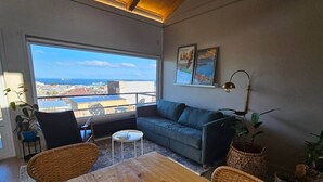 A modern living room with a large window, a sofa, a wooden table, and wicker chairs.