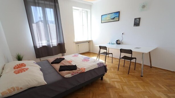 A bedroom with a bed, a desk, and a chair. There is a window with curtains, a radiator, and a small plant.