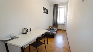 A small, clean room with a white table, a black chair, a bed with a gray and white patterned blanket, and a window with curtains.