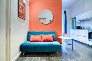 A room with a blue sofa, a round mirror on the wall, a small table with a book, and a kitchen area in the background.