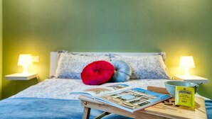 Ein Bett mit einer blauen Decke, einem roten und blauen Kissen, einer Zeitschrift, einer Tasse und einer Packung Twinings Zitronen-Aromatee auf einem Nachttisch.