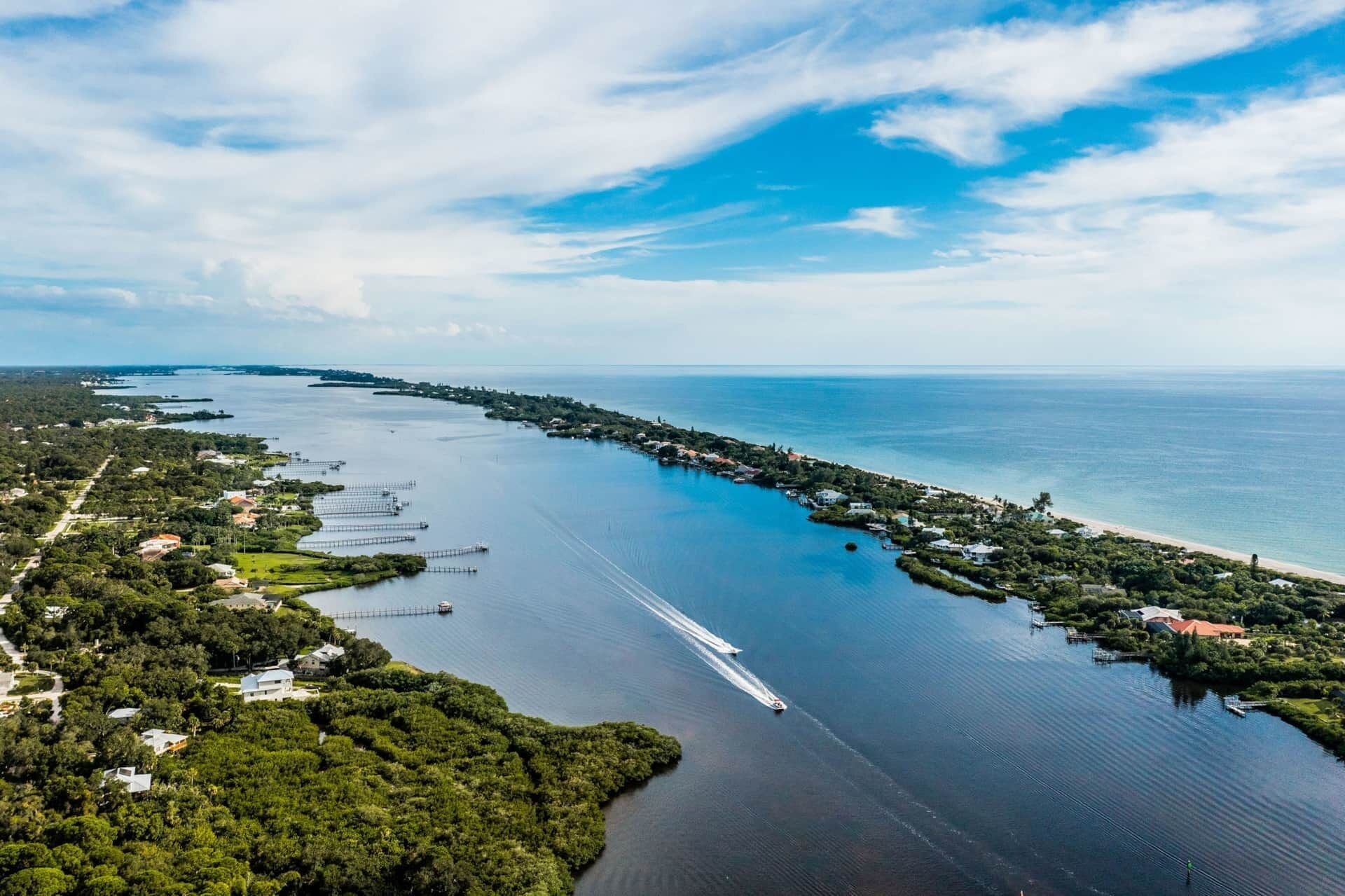 An aerial view of a coastal area with a long pier extending into the water, surrounded by residential buildings and greenery.