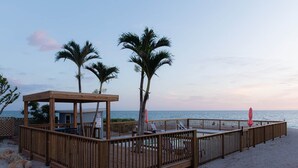 Eine Holzterrasse mit überdachter Sitzecke, Palmen und Blick auf das Meer.